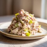 A mound of creamy ham salad mixed with diced celery, seasoned with pepper, served on a light plate.