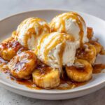 Three scoops of vanilla ice cream drizzled with caramel sauce over sautéed bananas in a dish of Bananas Foster.