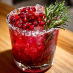 Close-up of a vibrant red cranberry pomegranate margarita with a salt rim, topped with fresh pomegranate seeds and rosemary.