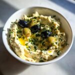 A white bowl filled with creamy dirty martini dip, garnished with whole green and black olives and fresh parsley.