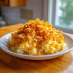 A scoop of creamy hashbrown casserole topped generously with golden, crunchy cornflakes served on a white plate.