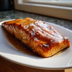 A perfectly cooked, glazed salmon fillet resting on a white plate with a sweet, savory sauce.