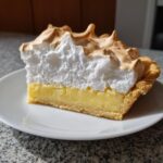 A tall slice of lemon meringue pie showing a thick, fluffy, toasted meringue topping over bright yellow lemon filling and a golden crust.