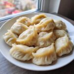 A white plate piled high with freshly steamed Traditional Chinese Dumplings, set near a window.