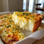 A serving spoon lifts a thick slice of cheesy baked mashed potatoes topped with melted cheddar and chives.