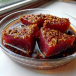 Three thick, deep-red tuna steak pieces soaking in a rich, dark MARINADE FOR TUNA STEAK topped with chili flakes and garlic.