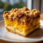 A close-up of a square slice of pumpkin crunch dessert with a thick, crumbly topping.