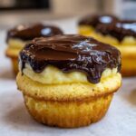A close-up of a delicious boston cream pie cupcake, featuring a split vanilla cake layer filled with pastry cream and topped with rich chocolate ganache.