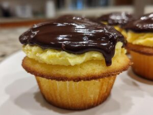 A close-up of a single boston cream pie cupcake featuring a vanilla cake base, thick pastry cream filling, and rich chocolate ganache topping.