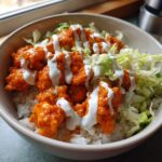 A close-up view of a delicious buffalo chicken bowl featuring orange buffalo-coated chicken pieces over white rice, topped with shredded lettuce and a drizzle of ranch dressing.