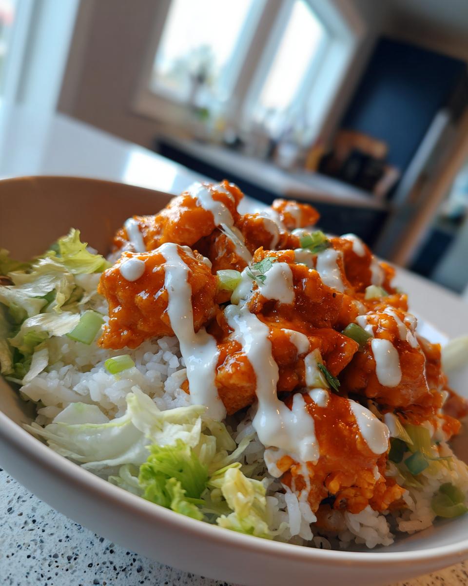 A close-up of a buffalo chicken bowl featuring orange sauced chicken, white rice, shredded lettuce, and a drizzle of ranch dressing.