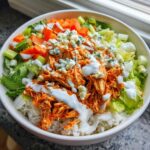 Close-up of a rice bowl featuring shredded buffalo chicken, lettuce, carrots, celery, blue cheese crumbles, and ranch dressing.