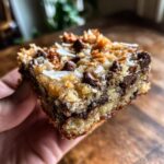 A hand holding a thick, chewy square of magic cookie bars topped with chocolate chips and coconut flakes.