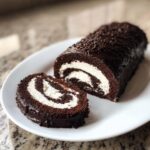 A rich chocolate yule log cake, partially sliced, revealing a white cream swirl filling.