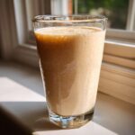 A tall glass filled with a thick, creamy banana smoothie, sitting on a sunlit windowsill.