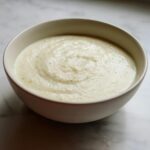 A close-up view of a bowl filled with smooth, creamy white cauliflower soup, lightly seasoned.