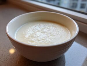 A close-up of a white bowl filled with smooth, creamy cauliflower soup, set near a window.