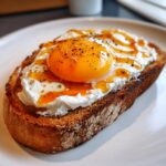 Close-up of creamy egg toast featuring a perfectly runny yolk, white cheese spread, and a drizzle of oil, seasoned with pepper.
