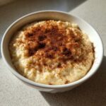A close-up of creamy rice pudding in a white bowl, generously dusted with ground cinnamon.