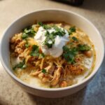 A close-up of a bowl of creamy crockpot white chicken chili topped with shredded cheese, sour cream, and fresh cilantro.