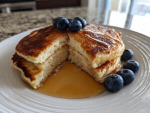 A stack of fluffy greek yogurt pancakes cut in half, drizzled with syrup and topped with fresh blueberries.