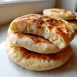 A stack of three golden-brown, fluffy pita bread rounds, with the top one broken open to show the airy interior.