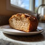 A thick slice of golden brown soda bread with a rustic crust resting on a white plate indoors.