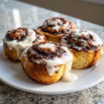 Four freshly baked cinnamon roll pastries topped with thick white icing, sitting on a white plate.