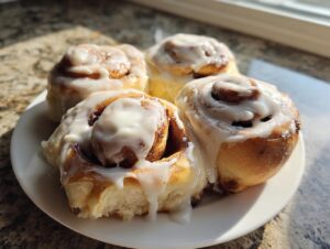 Four freshly baked cinnamon rolls generously covered in white icing, resting on a white plate.