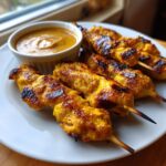 Close-up of perfectly grilled, golden chicken satay skewers served with a side of creamy peanut dipping sauce.