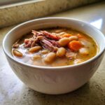 Close-up of a white bowl filled with rich Ham and Bean Soup, showing shredded ham, white beans, and sliced carrots in broth.
