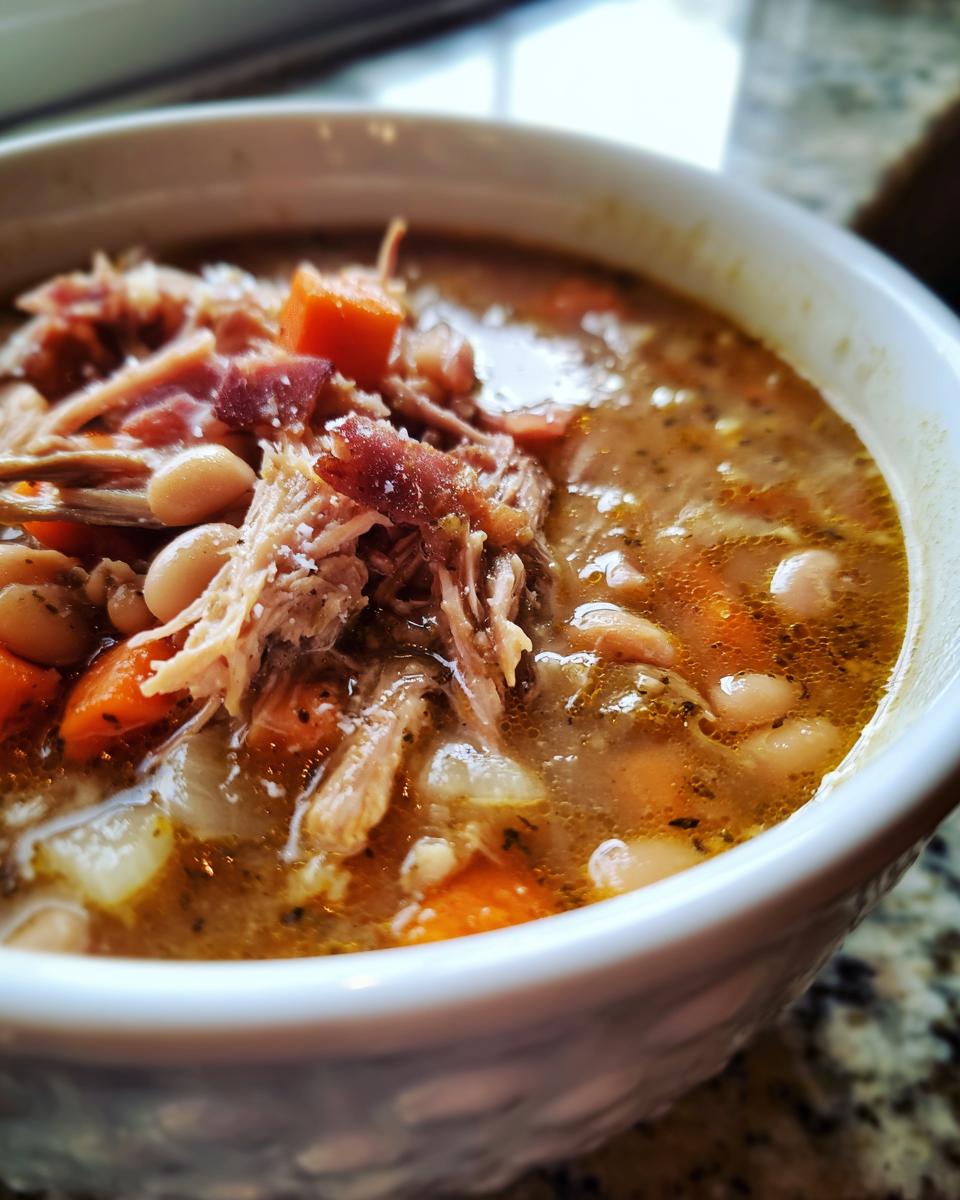 Close-up of a white bowl filled with rich Ham and Bean Soup, featuring shredded ham, white beans, and carrots.