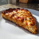 A thick slice of keto pizza crust topped with melted mozzarella, tomato sauce, and herbs, sitting on a white plate.