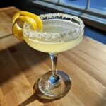 A bright yellow lemon drop cocktail served in a coupe glass with a sugar rim and lemon peel garnish.