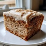 A square slice of moist applesauce cake topped with thick, creamy frosting and sprinkled with cinnamon.