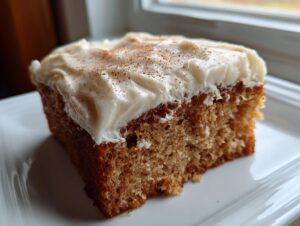 A close-up of a moist slice of applesauce cake topped with thick cream cheese frosting and a dusting of cinnamon.