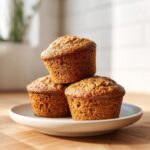 Three golden brown, moist bran muffins stacked on a small white plate, ready to eat.