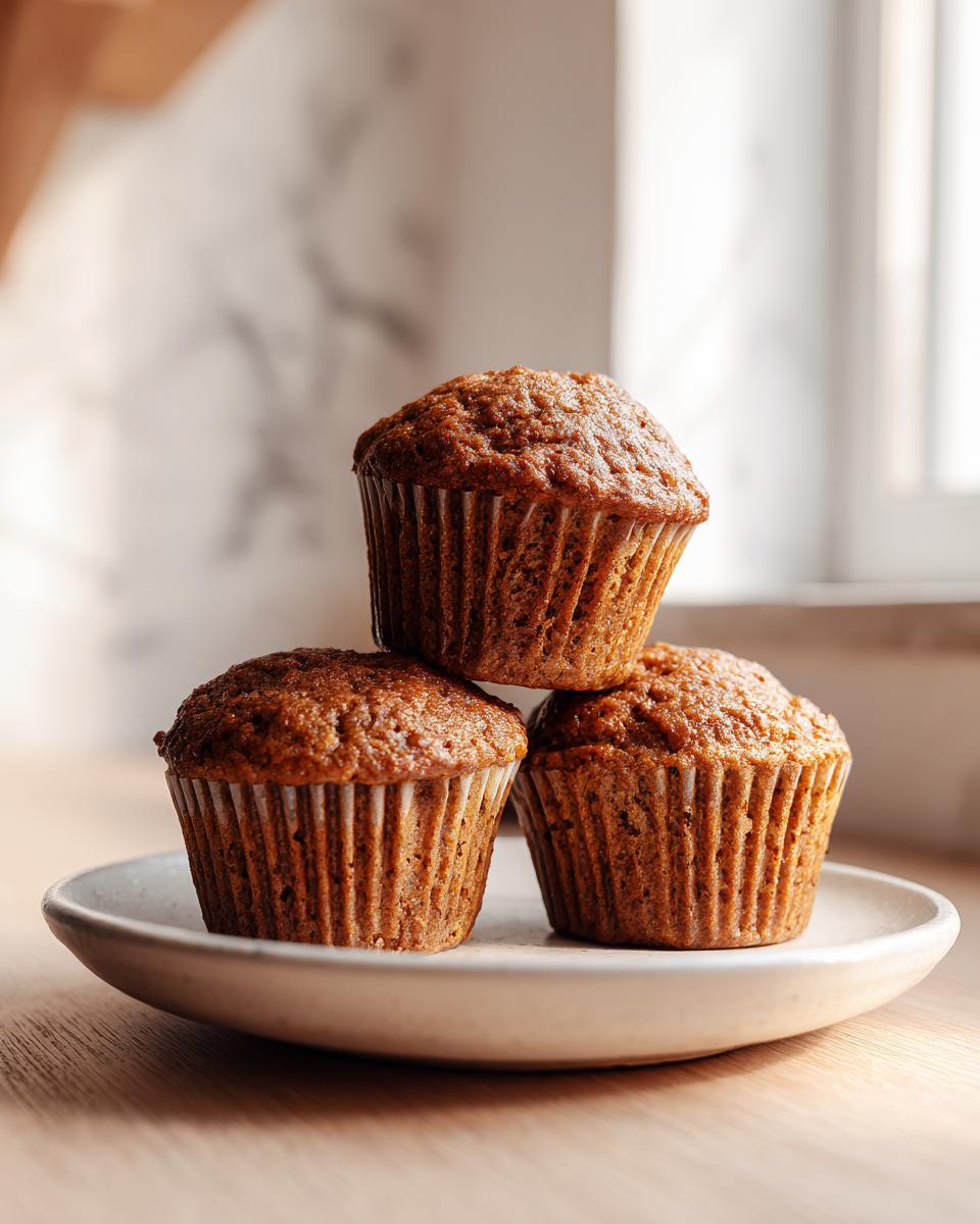 Three freshly baked, moist bran muffins stacked on a small, light-colored plate on a wooden surface.