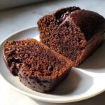 Two moist slices of dark, rich chocolate bread sitting on a white plate, highlighted by sunlight.