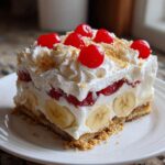 A single serving slice of no-bake banana split cake featuring layers of bananas, cream, cherries, and whipped topping.