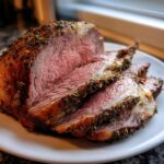 Close-up of a perfectly cooked, medium-rare rib roast sliced on a white plate, showing a thick herb crust.