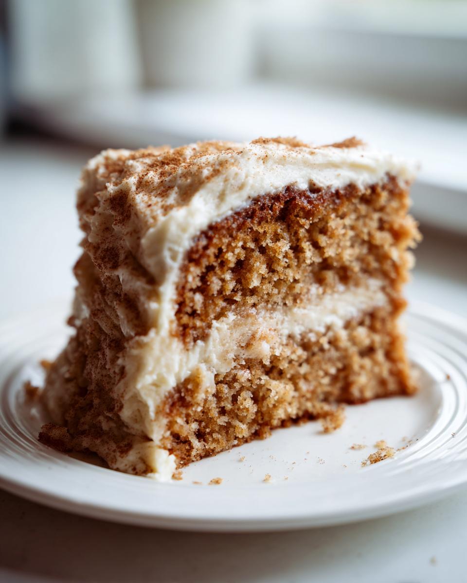 A tall slice of moist applesauce cake layered with cream cheese frosting and dusted with cinnamon.