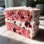 Close-up of a moist slice of pink cherry cake layered with white cream filling and visible whole cherries.