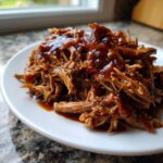 A close-up of tender, shredded Slow Cooker Pulled Pork generously covered in rich, glossy BBQ sauce on a white plate.