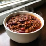 A steaming white bowl filled with rich, hearty crockpot chili featuring ground beef and kidney beans, set near a window.