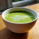 Close-up of a bowl of vibrant green asparagus soup with a light foam topping, sitting on a wooden table.
