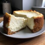 A thick slice of creamy white chocolate cheesecake next to the main cake, showing a golden graham cracker crust.
