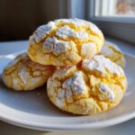 A stack of bright yellow lemon crinkle cookies generously coated in powdered sugar, sitting on a white plate.