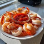 A white plate piled high with chilled, cooked shrimp surrounding a small glass bowl of bright red shrimp cocktail sauce.