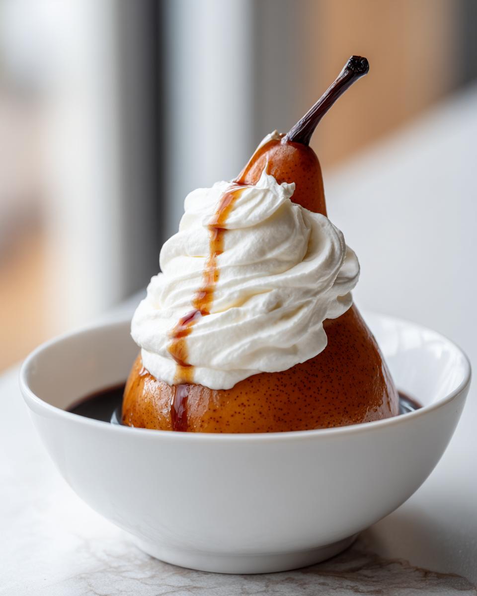 A single, beautifully poached pear topped with a swirl of whipped cream and caramel drizzle, served in a white bowl.